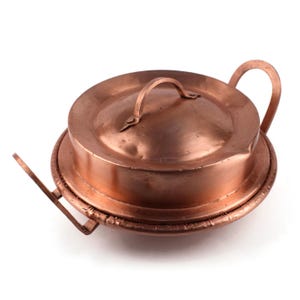Puede incluir: Un recipiente de cocina redondo de cobre con tapa a juego y dos asas laterales. La olla presenta un acabado metálico liso con un tono marrón rojizo cálido. La tapa tiene un asa central arqueada y la base un borde ancho y plano.