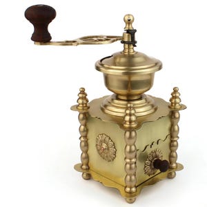 Può includere: Un'antica macinacaffè in ottone con manico e pomello in legno. Il macinacaffè presenta un motivo floreale decorativo sulla parte anteriore e sui lati, con dettagli ornati sulle gambe e sulla parte superiore. Il colore generale è oro caldo.