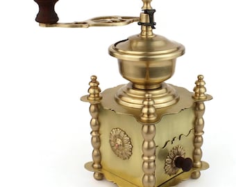 May include: A vintage brass coffee grinder with a wooden handle and knob. The grinder features a decorative floral design on the front and sides, with ornate detailing on the legs and top. The overall color is a warm gold.
