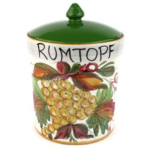 May include: A ceramic storage jar with a glossy green lid and a pointed finial. The white body features the hand-painted text RUMTOPF in black lettering, accompanied by a colorful illustration of yellow grapes and green leaves with red accents.
