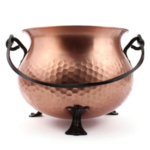 Può includere: Un calderone di rame con una texture martellata, manico nero e tre piedini decorativi. Il calderone ha una forma arrotondata e una finitura metallica lucida, ideale per uso decorativo.