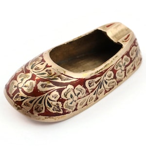 May include: A brass ashtray shaped like a shoe, adorned with detailed floral patterns in red and gold. The ashtray has a small, open interior for holding ashes and a decorative design around the edges.