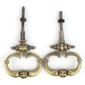 May include: Two brass drop pulls featuring a rounded, loop-shaped handle with a central decorative ribbed detail. Each pull includes a vertical mounting post with a threaded screw and a single hexagonal nut attached to the top section.