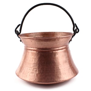 May include: A hammered copper pot with a wide, flared body and a smooth, rounded rim. The vessel features a dark, arched metal handle attached to the sides with looped brackets, creating a sturdy, functional design for this metallic container.