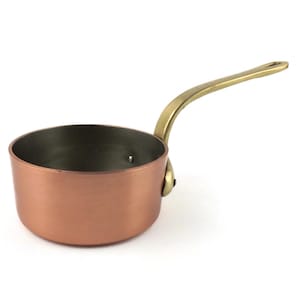 Puede incluir: Una cacerola de cobre con un asa de latón. La pequeña olla tiene un cuerpo redondeado y un asa larga y curvada. El interior de la sartén es de color oscuro, contrastando con el exterior de cobre.