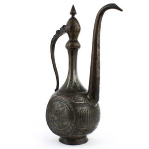 Op de afbeelding: Antieke zilverkleurige metalen kan met een lange, gebogen tuit en een handvat. De kan heeft ingewikkelde gegraveerde patronen en ontwerpen, waaronder cirkelvormige motieven op de afgeronde basis. De bovenkant heeft een puntige bekroning.