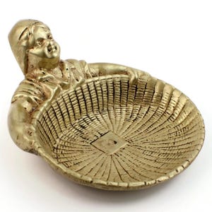 May include: A gold-colored figurine of a person holding a basket. The basket is oval-shaped with a woven texture. The person is depicted with a smiling expression and is wearing a hooded garment.