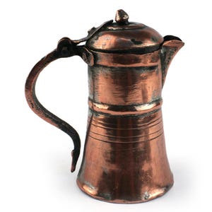 Puede incluir: Una jarra de cobre desgastada con tapa abatible, asa curva y un pequeño pico vertedor. La superficie metálica muestra signos de oxidación y desgaste, con estrías horizontales alrededor de la base y una pátina oscura y texturizada.