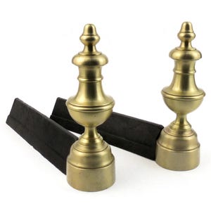 May include: Two antique brass fireplace andirons with a classic urn design. The andirons have a gold finish and are paired with two black, rectangular firebacks. The andirons are approximately 25 cm tall.