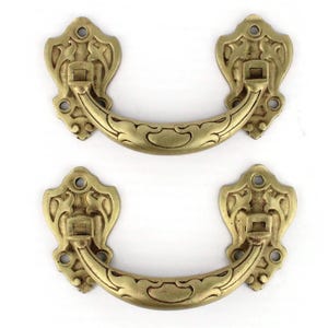 Könnte beinhalten: Zwei antike Schubladengriffe aus Messing mit aufwendigen, floral inspirierten Designs. Jeder Griff verfügt über einen gebogenen Griff und dekorative Rückplatten mit aufwendigen Details. Die Hardware hat eine warme, goldene Farbe, die auf eine gealterte Oberfläche hindeutet und für die Möbelrestaurierung geeignet ist.