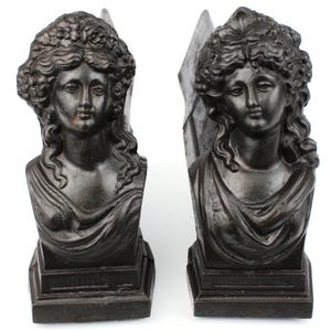 Pode incluir: Par de suportes para livros em bronze escuro com bustos femininos clássicos com cabelos detalhados e roupas drapeadas. Cada busto é montado em uma base retangular, criando um item decorativo.