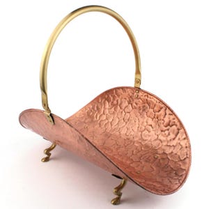May include: A copper-coloured magazine rack with a hammered texture and brass handle and feet. The rack has a curved design and is supported by three decorative feet. The handle arches over the top.