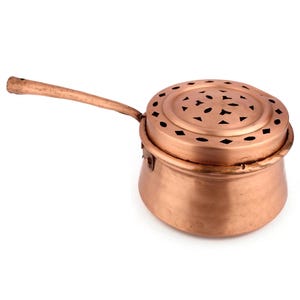 Puede incluir: Una olla de cobre con una tapa que presenta un patrón decorativo de formas recortadas. La olla tiene un asa larga y es ideal para cocinar o servir.