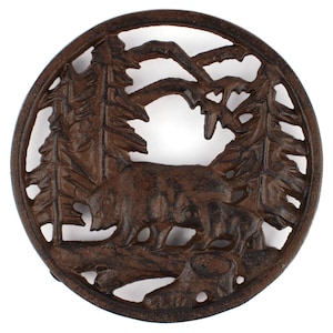 Peut inclure: Un dessous de plat circulaire en fonte marron avec un motif ajouré représentant un ours et son petit marchant dans une forêt de pins, avec des montagnes en arrière-plan. La surface métallique présente une finition texturée aux tons brun foncé.