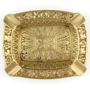 May include: An antique brass ashtray with a rectangular shape and detailed floral and leaf relief patterns. The ashtray has four cigarette rests. The edges are decorated with flowers, and the centre features an intricate leaf pattern.