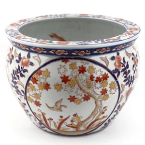 May include: A round, white ceramic planter with a detailed floral and tree design. The planter features a blue rim with floral patterns and a central motif of a tree with red and yellow leaves and a flying bird.