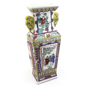 May include: A white porcelain vase with a rectangular shape and hand-painted scenes. The vase features yellow handles and is decorated with red, blue, and green accents. The vase is a decorative item, likely for display.
