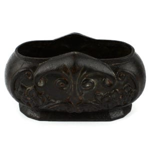 Peut inclure: Un pot de fleurs ovale en fonte brun foncé avec une surface texturée. L'extérieur présente des détails en relief, notamment des volutes symétriques et des motifs floraux sur les côtés, avec une base solide et un dessus ouvert pour le rangement.
