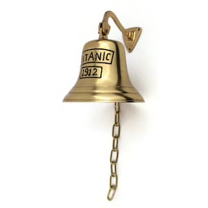 May include: A gold-colored brass ship's bell with a black inscription that reads "TITANIC 1912". The bell is attached to a wall mount and has a chain hanging from the bottom.