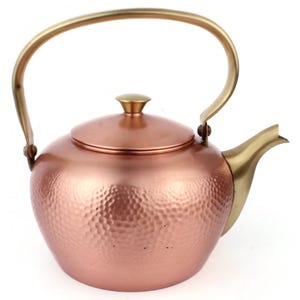 May include: A copper tea kettle with a hammered texture, featuring a brass handle and spout. The lid is topped with a small brass knob. The kettle has a rounded body and a classic design, suitable for brewing tea.