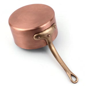 Puede incluir: Una cacerola de cobre con un asa de latón. La olla tiene un cuerpo redondeado y un asa larga con un lazo en el extremo. El cobre tiene un acabado brillante y el asa es de color dorado.