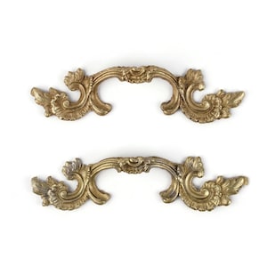 May include: Two matching gold-toned metal drawer pulls featuring an ornate, symmetrical design with intricate scrollwork and leaf-like patterns extending from the center handle to the decorative ends on both sides of each piece.