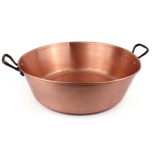 May include: A copper-coloured, round pan with two dark-coloured handles. The pan has a wide, shallow bowl shape, suitable for cooking or serving. The copper surface reflects light, giving it a shiny appearance. The handles are curved and attached to the sides.
