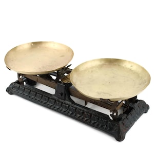 Peut inclure: Une ancienne balance de précision avec deux plateaux ronds en laiton. La balance a une base noire ornée et un cadre en métal foncé. Les plateaux sont vides, sur fond blanc.