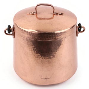 Pode incluir: Uma panela de cobre com tampa e alça. A panela tem uma textura martelada e duas alças laterais. A tampa tem uma pequena alça. A panela é de uma cor marrom-avermelhada quente.