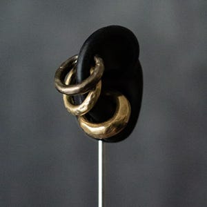 May include: Three gold-toned hoop earrings displayed on a black earring holder. The earrings have a textured, hammered appearance. The background is a solid, muted gray.