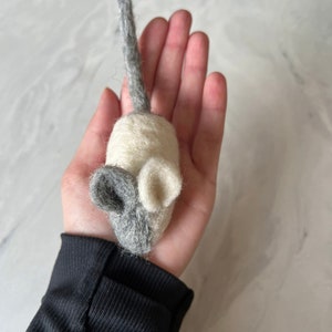 Puede incluir: Un pequeño juguete de ratón de fieltro hecho a mano con cuerpo blanco y orejas y cola grises.