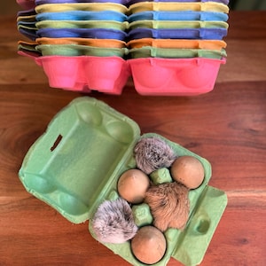 May include: A green cardboard egg carton with five brown wooden eggs and two gray and brown fuzzy toy chicks inside