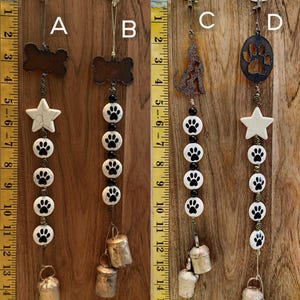May include: Four decorative dog-themed wind chimes. Each chime features a different top design: a bone, a star, a wolf, and a paw print. The chimes have paw print beads, small bells, and are suspended from a white hook. The background is a wooden surface.