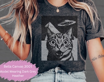 May include: A dark grey heather t-shirt with a black and white illustration of a cat with a surprised expression. The cat is looking up at a flying saucer. The text "Bella Canvas 3001" is visible in the bottom left corner of the image.