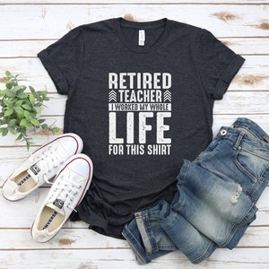 Puede incluir: Una camiseta gris oscuro con el texto "Retired Teacher I worked my whole life for this shirt" en blanco. La camiseta est&aacute; colocada sobre una superficie de madera blanca con un par de zapatos de lona blancos y unos vaqueros azules.