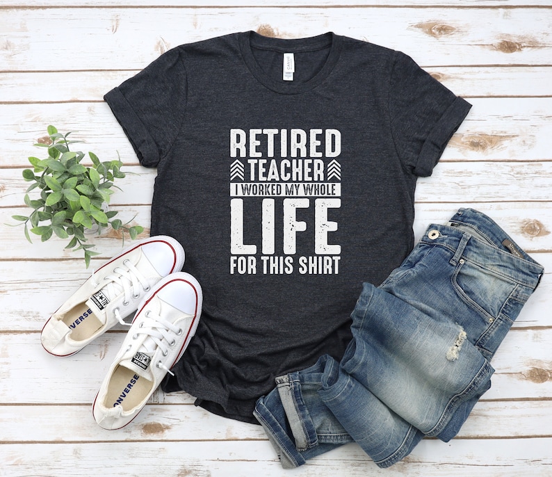 Puede incluir: Una camiseta gris oscuro con el texto "Retired Teacher I worked my whole life for this shirt" en blanco. La camiseta est&aacute; colocada sobre una superficie de madera blanca con un par de zapatos de lona blancos y unos vaqueros azules.