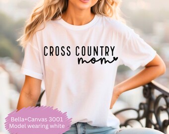 Może przedstawiać: Biała koszulka z czarnym napisem "Cross Country Mom" z symbolem serca. Modelka nosi koszulkę i niebieskie dżinsy.