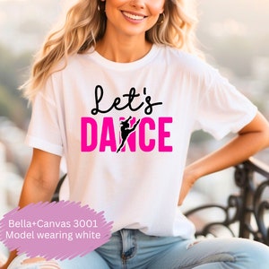 Peut inclure: Un t-shirt blanc avec le texte "Let's Dance" en lettres roses et une silhouette noire d'une danseuse. Le mannequin porte le t-shirt et est assis sur une balustrade.