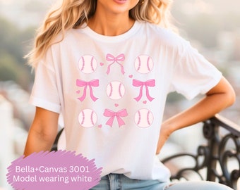 May include: White t-shirt featuring a repeating pattern of pink baseballs and bows. The baseballs have pink stitching and are interspersed with pink bows and small pink hearts. The text "Bella+Canvas 3001 Model wearing white" is at the bottom.