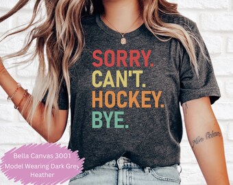 May include: A dark grey t-shirt with the text "SORRY. CAN'T. HOCKEY. BYE." printed in rainbow colors. The model is wearing the shirt with blue jeans.