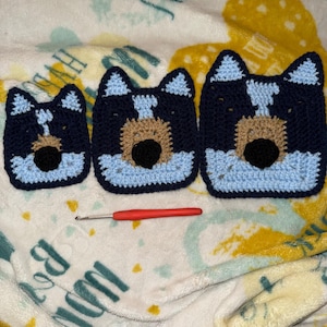 May include: Three crocheted dog faces in blue, brown, and white. The faces are different sizes and are arranged in a row on a white and yellow blanket. A red crochet hook is in front of the faces.
