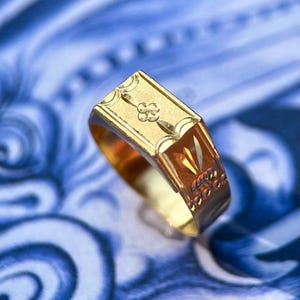 K&ouml;nnte beinhalten: Ein Goldring mit einer rechteckigen Kopfplatte, die ein eingraviertes vierbl&auml;ttriges Blumenmotiv sowie dekorative geometrische Muster an den Seiten aufweist. Der Ring hat eine polierte Oberfl&auml;che und eine flache Oberseite.