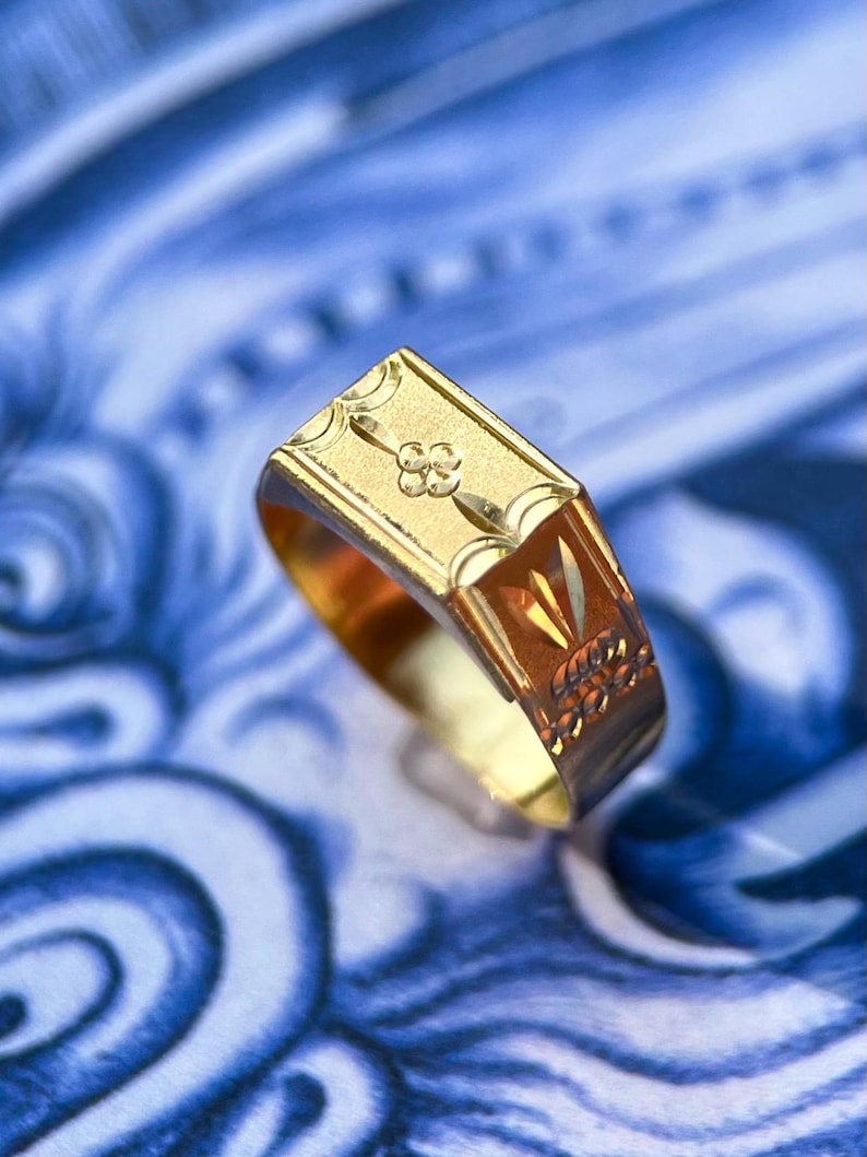 K&ouml;nnte beinhalten: Ein Goldring mit einer rechteckigen Kopfplatte, die ein eingraviertes vierbl&auml;ttriges Blumenmotiv sowie dekorative geometrische Muster an den Seiten aufweist. Der Ring hat eine polierte Oberfl&auml;che und eine flache Oberseite.