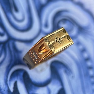 K&ouml;nnte beinhalten: Ein Goldring mit einer rechteckigen Vorderseite mit strukturierter Oberfl&auml;che, einem kleinen eingravierten Blumenmotiv und einem geometrischen Muster an der Seite. Der Ring hat eine polierte Oberfl&auml;che und zeigt feine Details am oberen Teil.