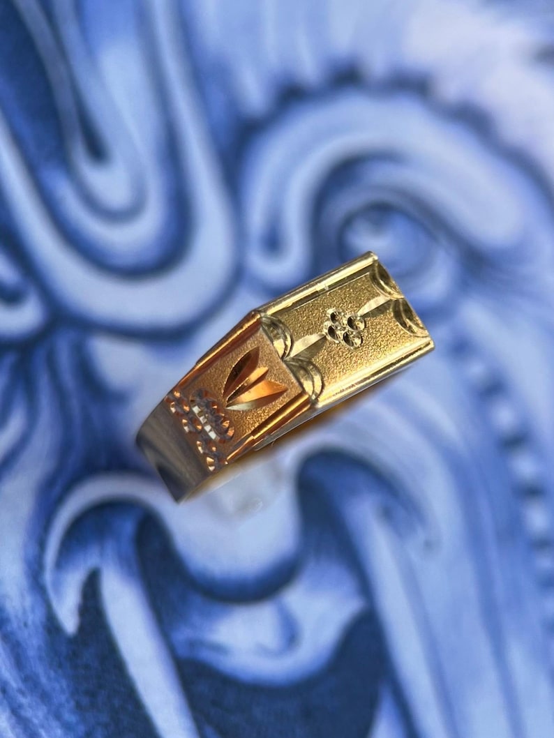 K&ouml;nnte beinhalten: Ein Goldring mit einer rechteckigen Vorderseite mit strukturierter Oberfl&auml;che, einem kleinen eingravierten Blumenmotiv und einem geometrischen Muster an der Seite. Der Ring hat eine polierte Oberfl&auml;che und zeigt feine Details am oberen Teil.