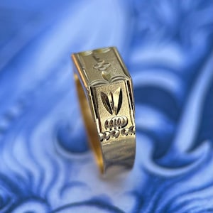K&ouml;nnte beinhalten: Ein goldener Ring mit einer rechteckigen Oberseite und komplexen Gravurmustern. Das Design umfasst ein zentrales blumenartiges Motiv, ein spitzes blattf&ouml;rmiges Element und eine Reihe kleiner kreisf&ouml;rmiger Vertiefungen am unteren Rand.