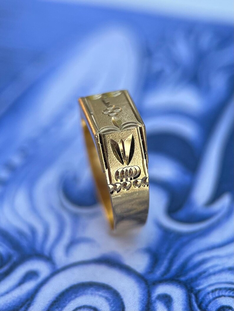 K&ouml;nnte beinhalten: Ein goldener Ring mit einer rechteckigen Oberseite und komplexen Gravurmustern. Das Design umfasst ein zentrales blumenartiges Motiv, ein spitzes blattf&ouml;rmiges Element und eine Reihe kleiner kreisf&ouml;rmiger Vertiefungen am unteren Rand.