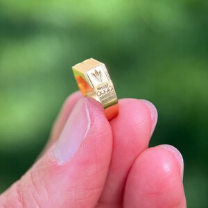 K&ouml;nnte beinhalten: Ein goldener Ring mit einer flachen, rechteckigen Oberseite, die ein eingraviertes Blumenmotiv und eine Reihe kleiner kreisf&ouml;rmiger Vertiefungen an der Seite aufweist. Der Ring hat eine polierte Oberfl&auml;che und eine geometrische Form.