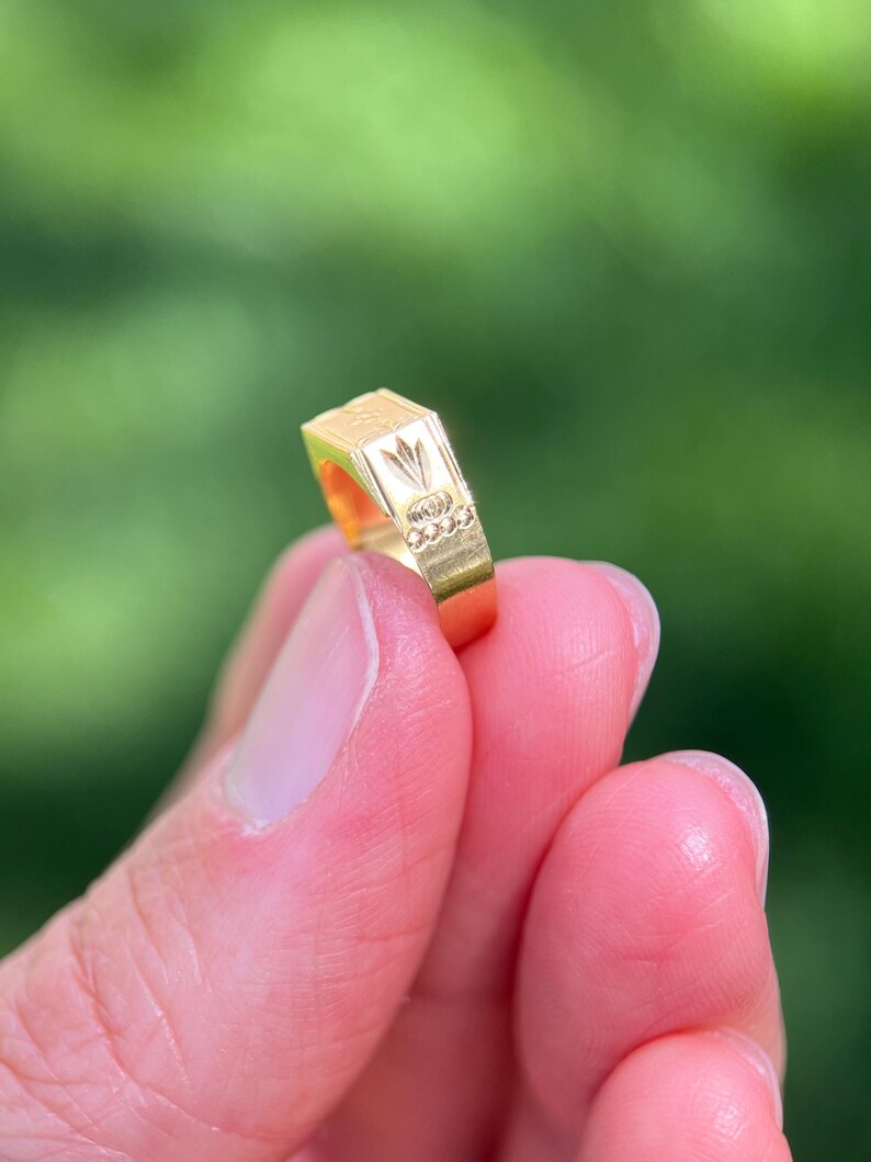 K&ouml;nnte beinhalten: Ein goldener Ring mit einer flachen, rechteckigen Oberseite, die ein eingraviertes Blumenmotiv und eine Reihe kleiner kreisf&ouml;rmiger Vertiefungen an der Seite aufweist. Der Ring hat eine polierte Oberfl&auml;che und eine geometrische Form.