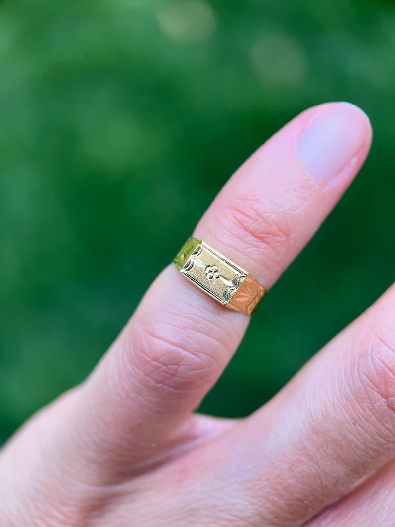 K&ouml;nnte beinhalten: Ein goldener Siegelring mit einer rechteckigen Platte, die in der Mitte ein eingraviertes Blumenmotiv und an den Seiten dekorative &Auml;tzmuster aufweist. Der Ring hat eine polierte Oberfl&auml;che und sitzt an einem Finger, was die Metallarbeit zeigt.